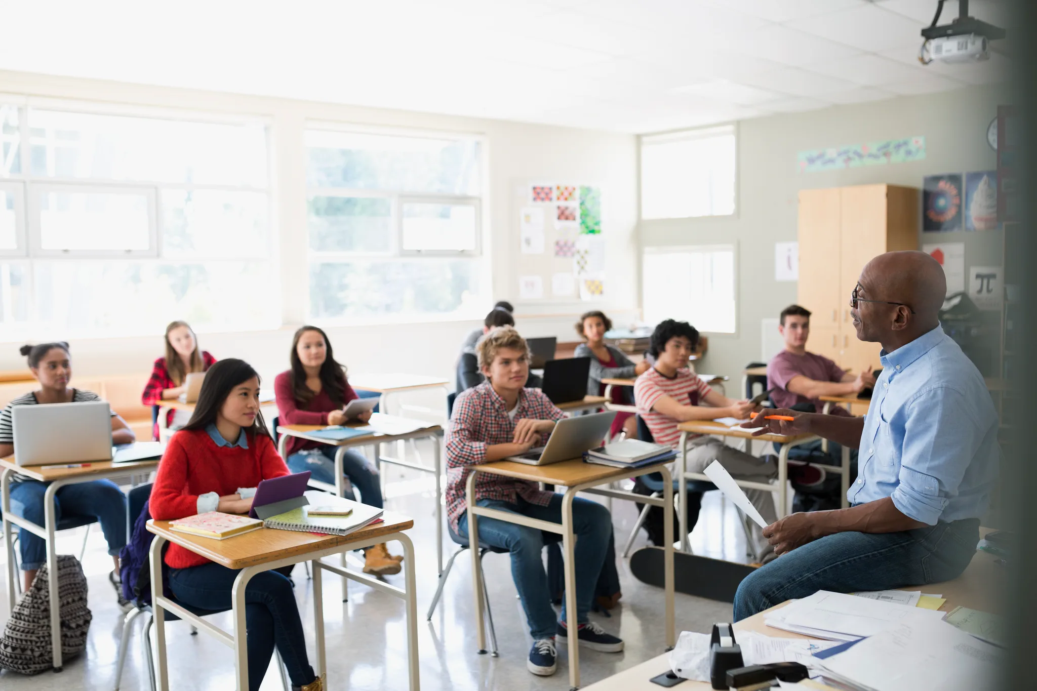 A teacher talking to high school students seated at desks in a bright classroom