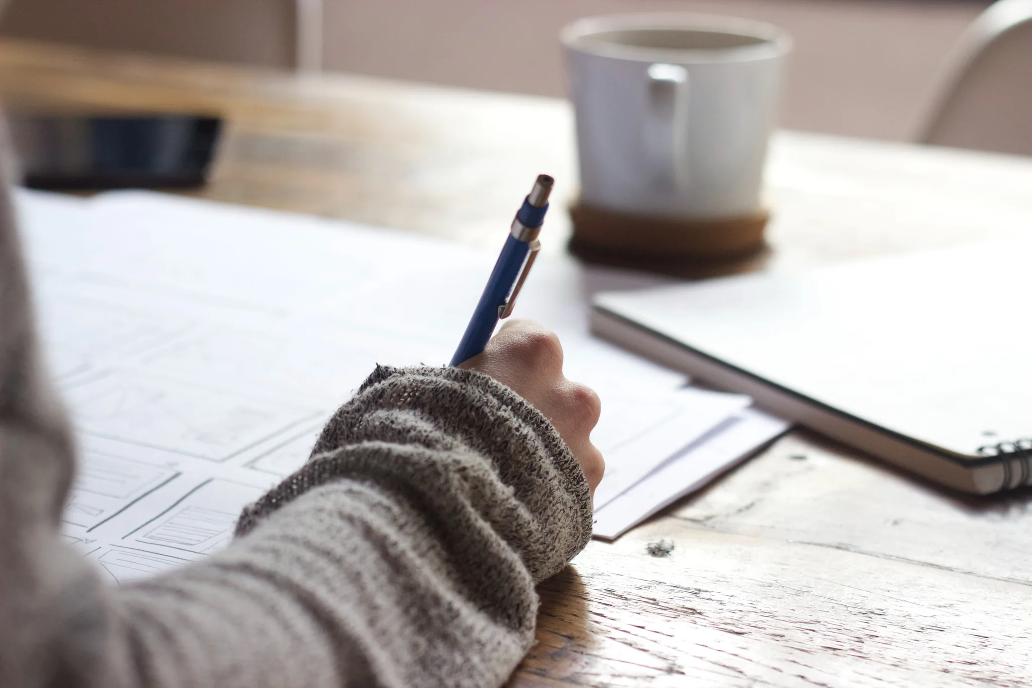 A person writing on paper with a pen beside a mug of coffee